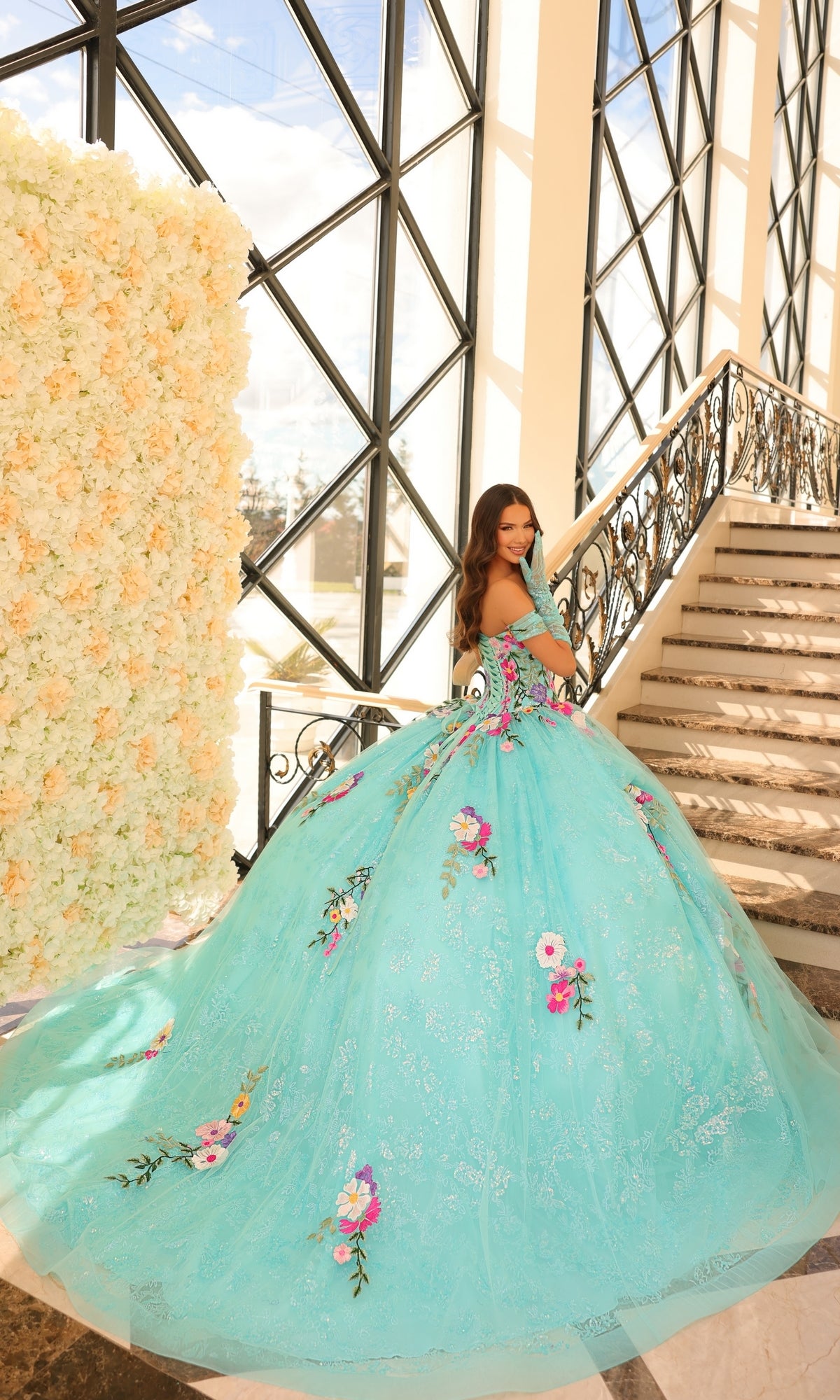 A woman wears the Quinceanera Dress 54200 By Amarra, featuring floral embroidery and an off-the-shoulder design, while standing by a staircase and a wall of white flowers in a sunlit room with large windows.
